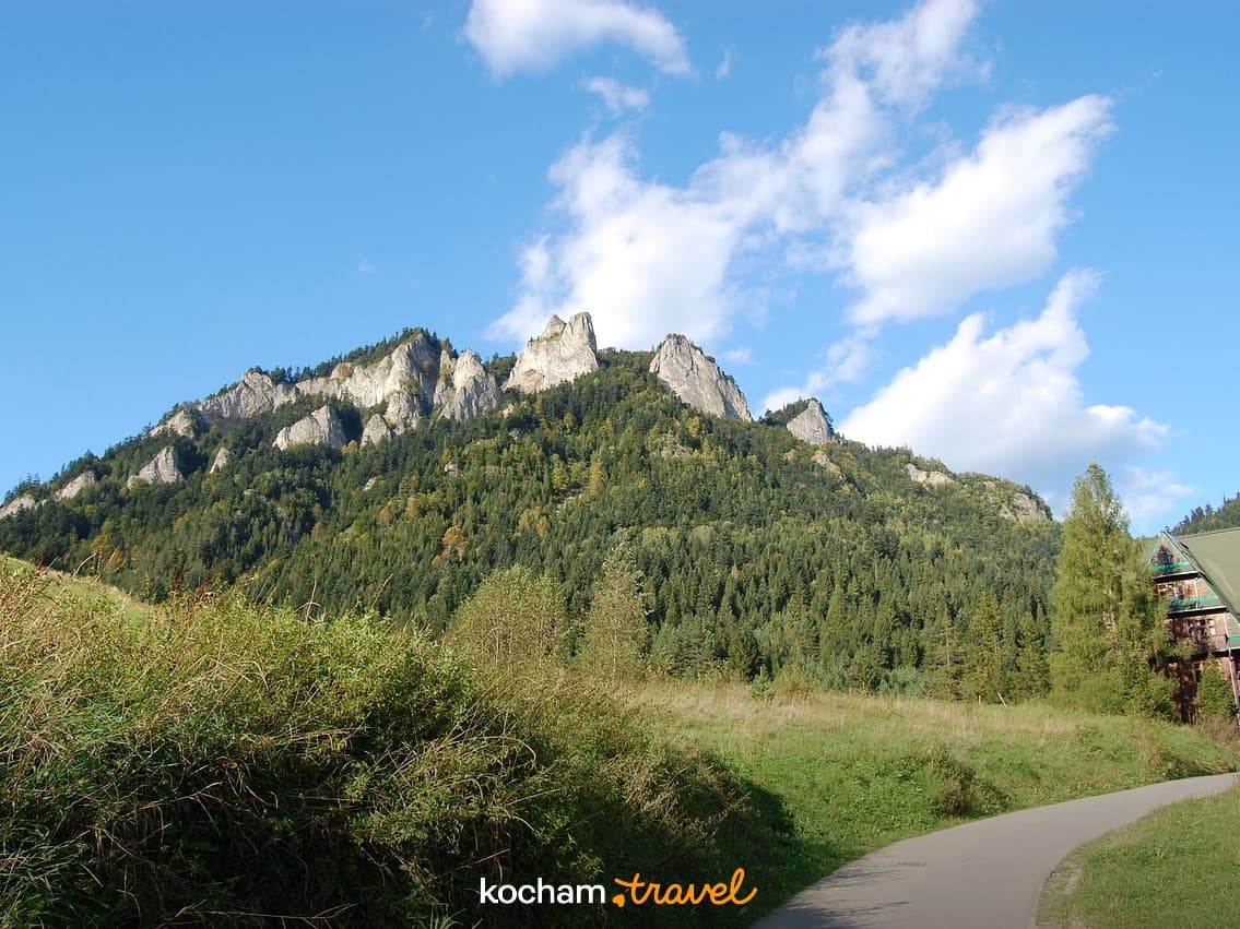 Słoneczny górski krajobraz, skaliste szczyty, zielony las, kręta droga, budynek.