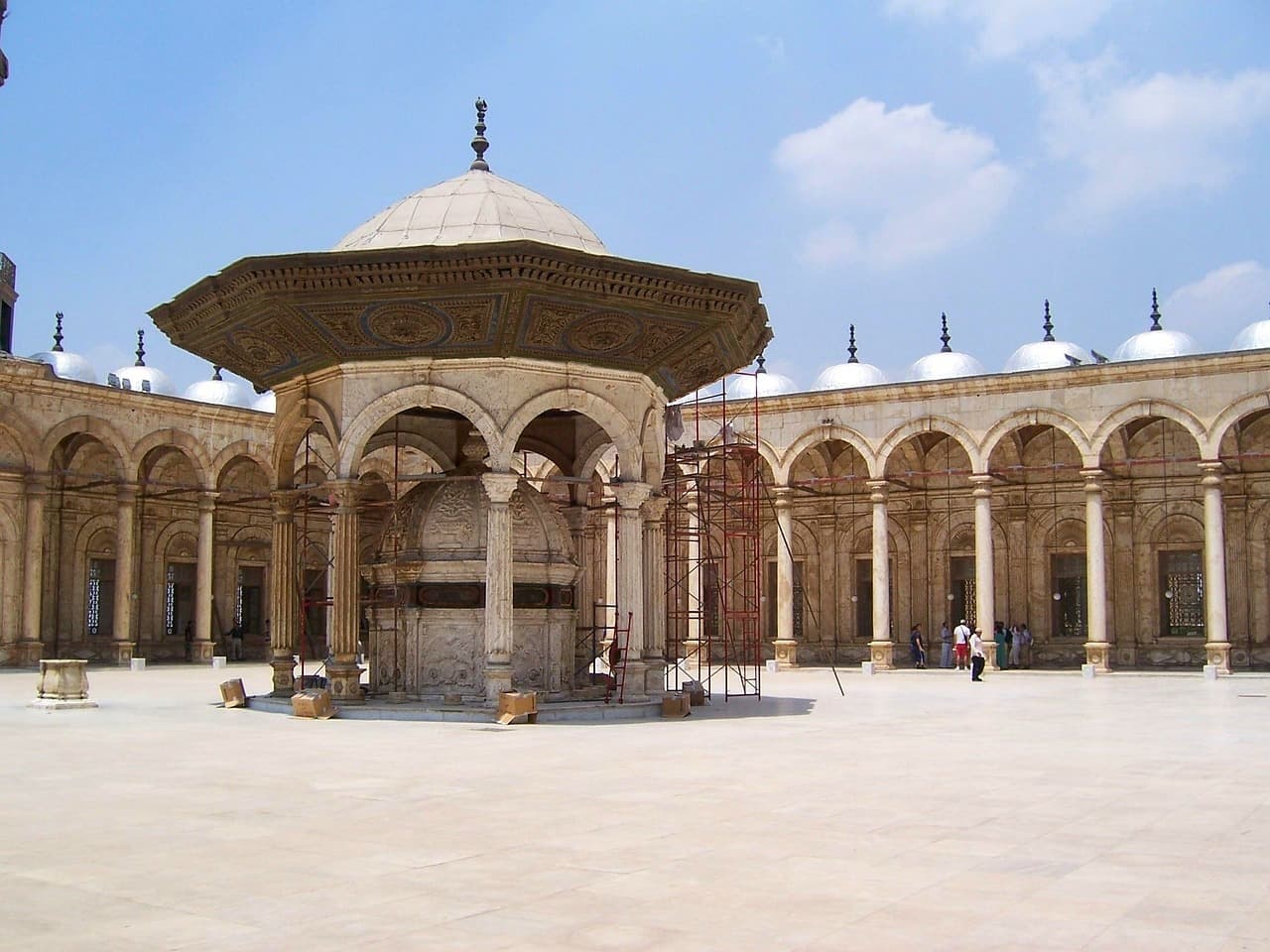 Słoneczny dziedziniec meczetu z ozdobną fontanną i arkadowymi kolumnadami.