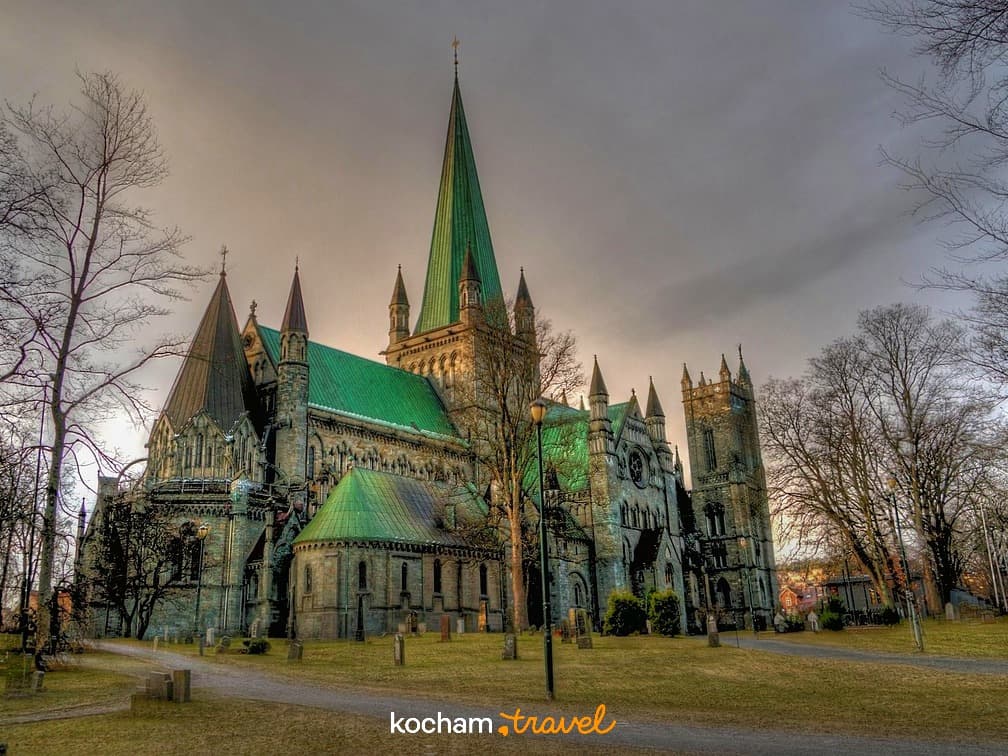 Gotycka katedra z zielonymi dachami, nagrobki, drzewa, pochmurne niebo.