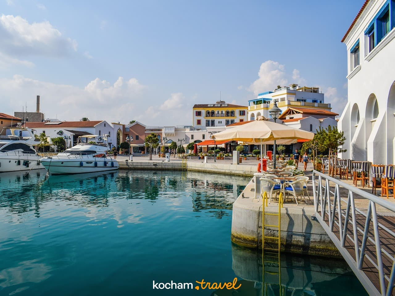Słoneczna marina z jachtami i restauracjami nad turkusową wodą.