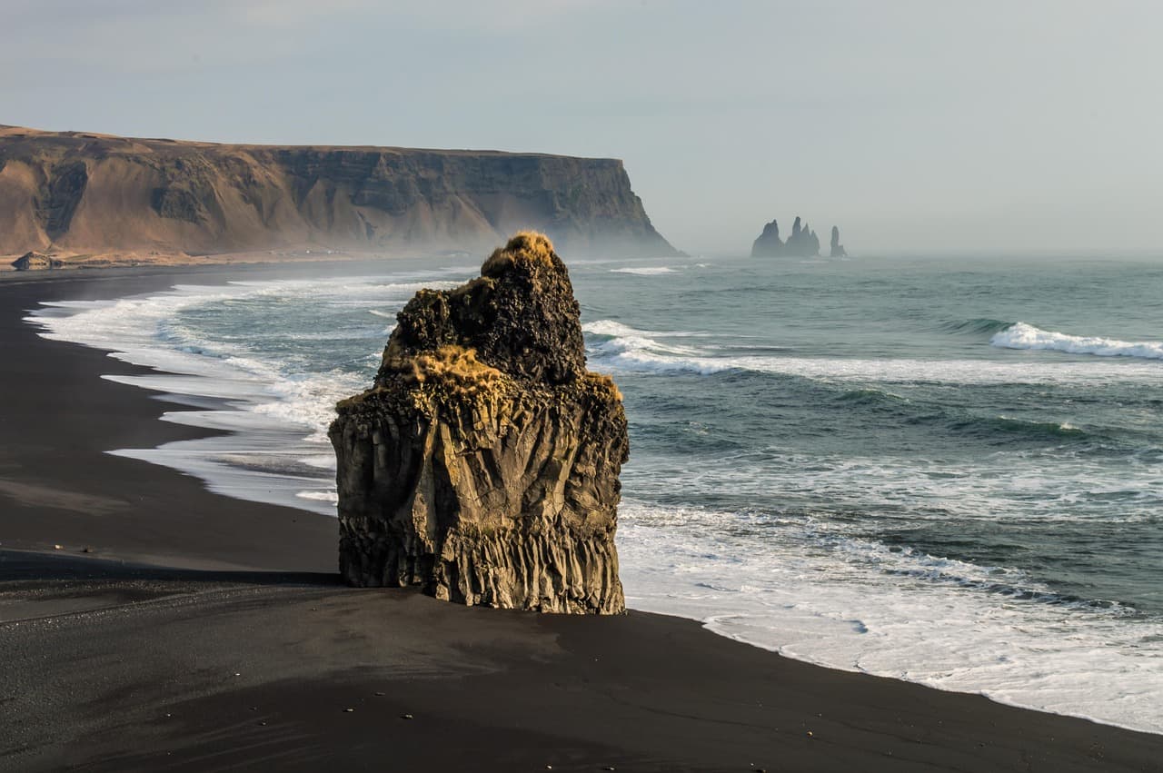 Samotna skalna formacja na czarnej plaży, fale oceanu, klify.