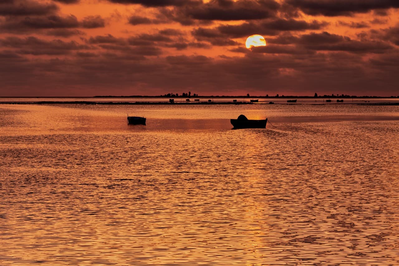 Dramatyczny zachód słońca nad morzem, pomarańczowe niebo, sylwetki łodzi.