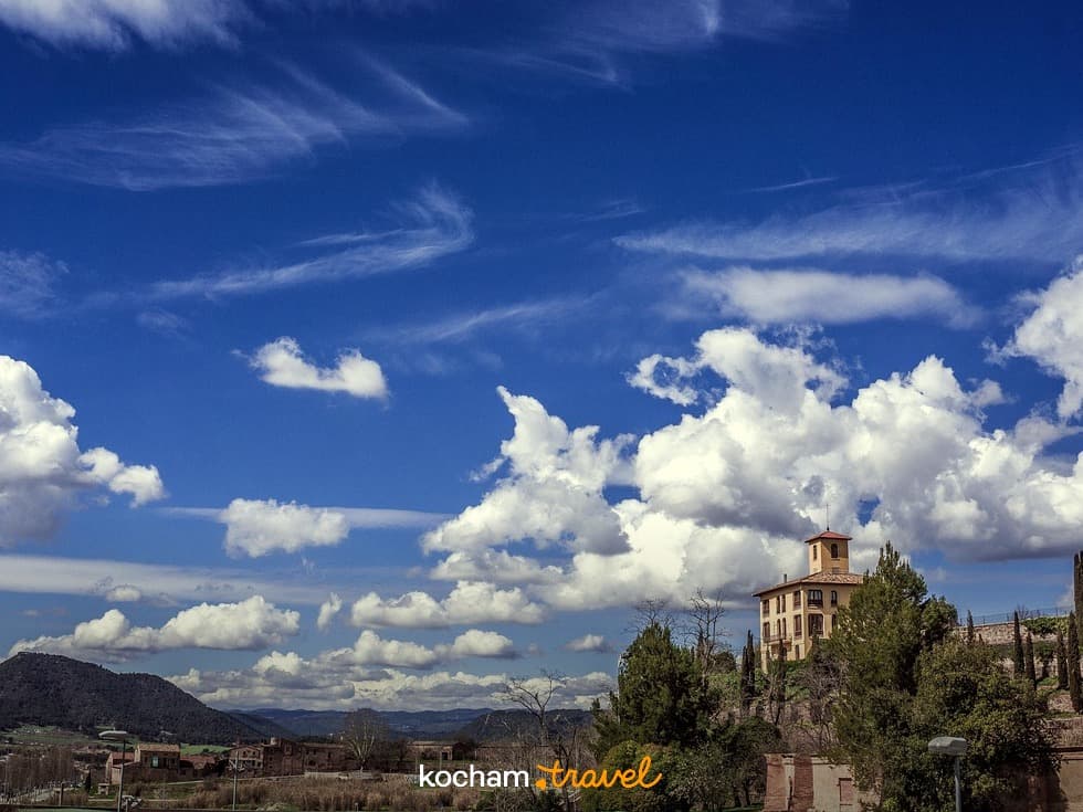 Historyczny budynek na wzgórzu, niebieskie niebo, białe chmury, zielone drzewa, góry.
