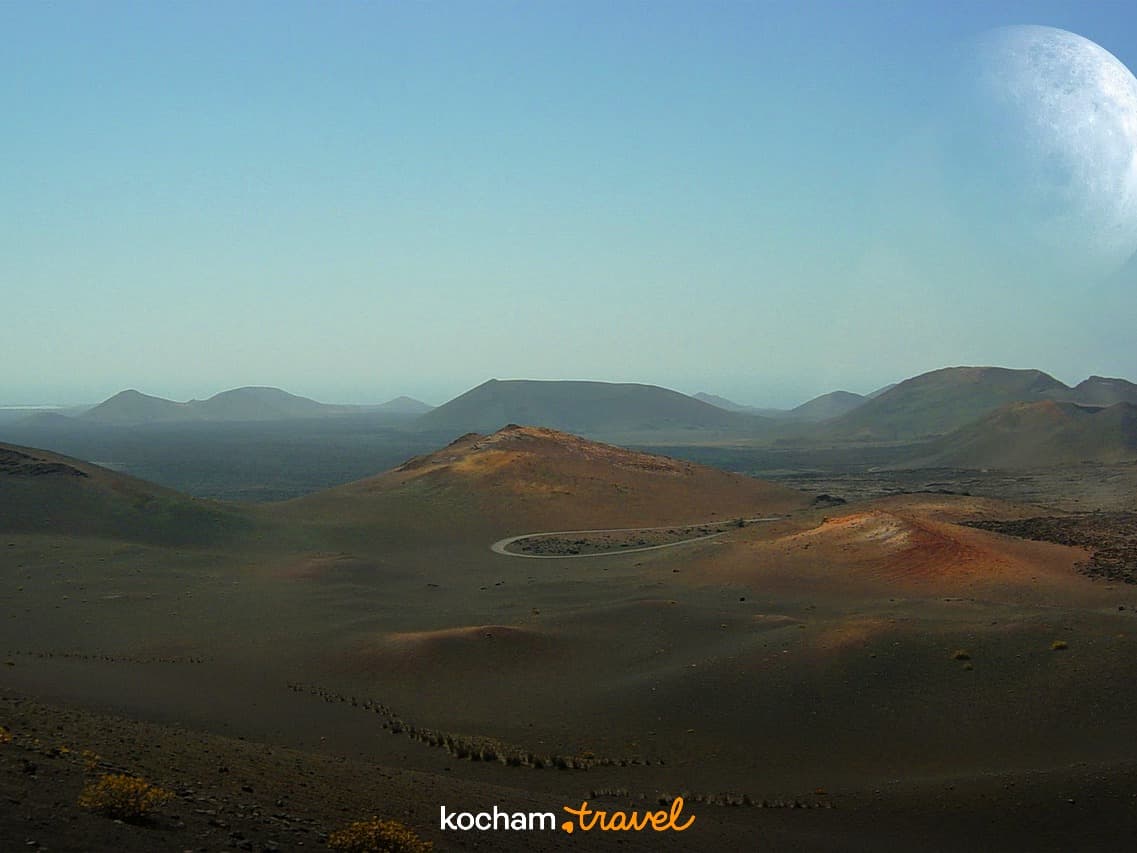 Rozległy krajobraz wulkaniczny z księżycem, ciemnymi wzgórzami, krętą drogą.